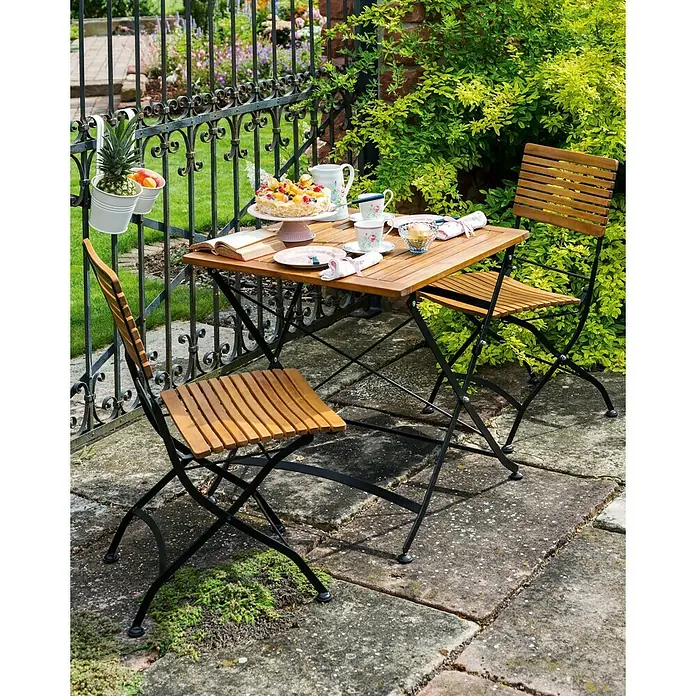 Sunfun Moni Balkonski stol - 75 x 75 cm, Akacija - Vanjski prizor koji prikazuje drveni stol i stolice postavljene s posuđem i hranom, nasuprot vrtne pozadine s bujnim zelenilom.
