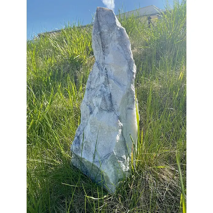 Gartendeko Monolithe - Marmor, Maße Stein: H: 40 - 55 cm - Nahaufnahme eines großen weißen Calcitkristalls, der auf einem grasbewachsenen Hügel steht. Das Sonnenlicht betont seine Textur.