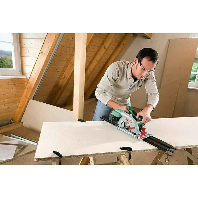 Bosch Handkreissäge PKS 55 A - 1.200 W, Ø 160 mm, 5.600 U/min - Ein Mann benutzt eine Bosch-Kreissäge mit Führungsschiene, um ein Brett zu schneiden, während er Sicherheitsbrille trägt und in einem Dachgeschoss arbeitet.