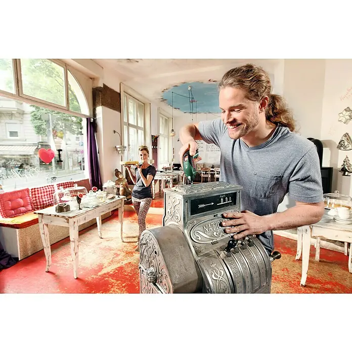 Bosch Akkuschrauber IXO V - 3,6 V, 1 Akku, 1,5 Ah, Leerlaufdrehzahl: 0 U/min - 215 U/min, 10 -tlg. - Ein Mann repariert lächelnd eine antike Registrierkasse in einem charmanten Café, während eine Frau im Hintergrund Kuchen serviert.
