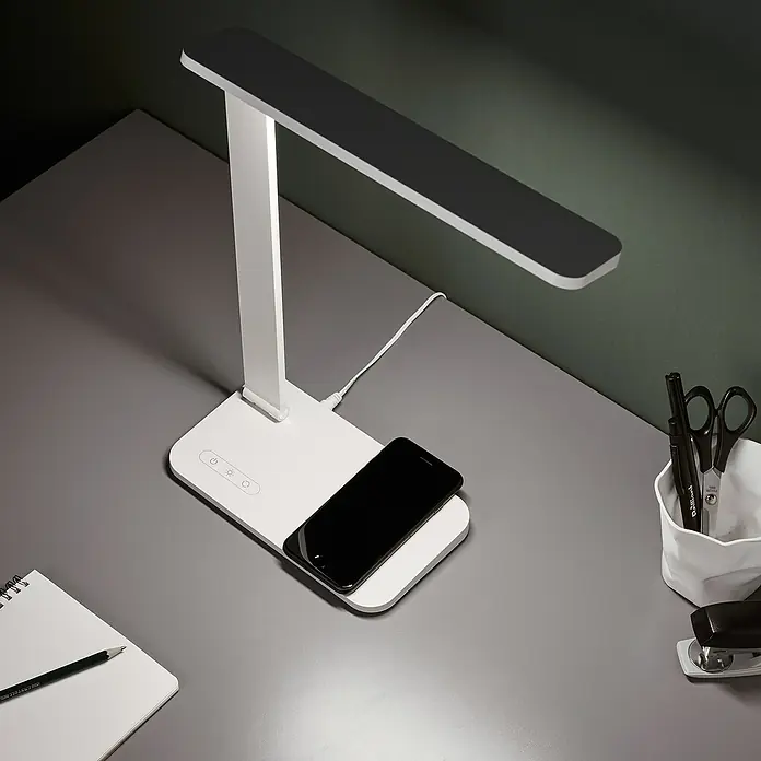 LED-Tischleuchte Seamont - 9 W, L x B x H: 31 x 12 x 37,4 cm, Weiß - Eine moderne Schreibtischlampe mit kabelloser Ladestation für Smartphones auf einem grauen Schreibtisch.