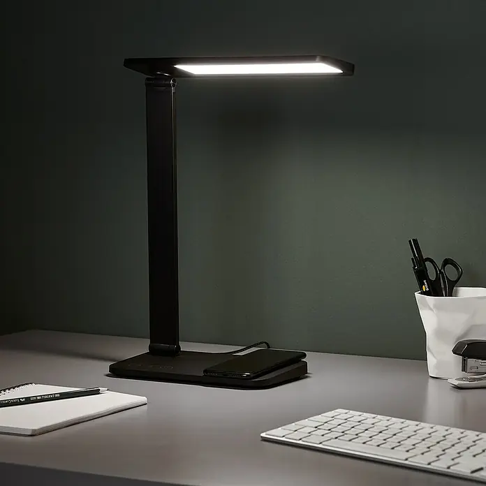 LED-Tischleuchte Seamont - 9 W, L x B x H: 31 x 12 x 37,4 cm, Schwarz - Eine moderne, schwarze Schreibtischlampe mit LED-Beleuchtung und kabelloser Ladefunktion für Smartphones auf einem grauen Schreibtisch.