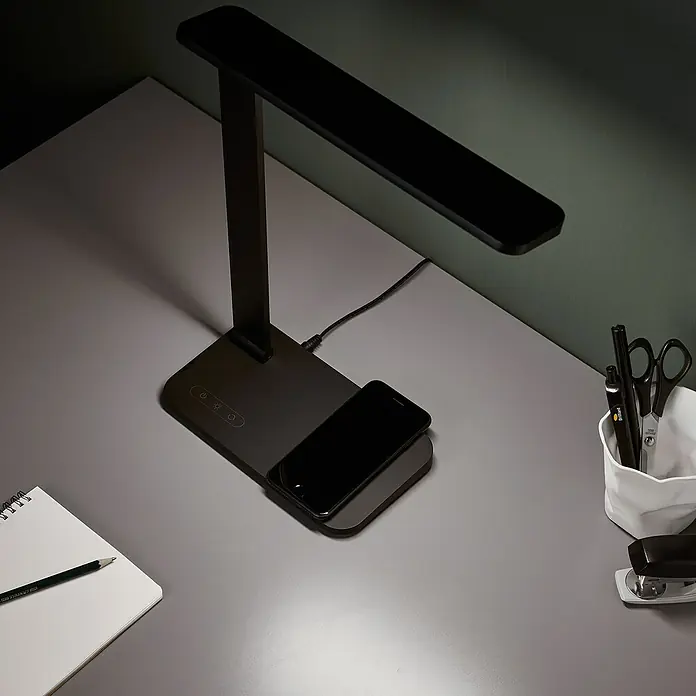 LED-Tischleuchte Seamont - 9 W, L x B x H: 31 x 12 x 37,4 cm, Schwarz - Ein moderner schwarzer Schreibtischlampe mit kabelloser Ladestation für ein Smartphone auf einem grauen Schreibtisch mit Büromaterial.
