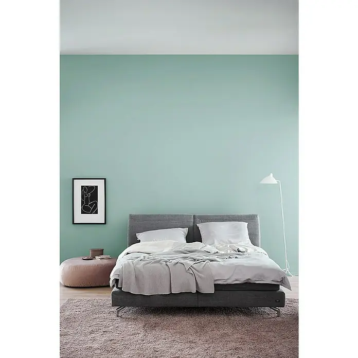 SCHÖNER WOHNEN-Farbe Wandfarbe Trendfarbe - Maui, 2,5 l, Matt, Konservierungsmittelfrei - Ein graues Boxspringbett mit weichen, weißen Kissen und einer grauen Strickdecke auf einem flauschigen Teppich vor einer mintgrünen Wand.