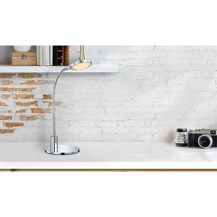 Globo LED-Tischleuchte Pegasi - 3 W, Chrom, Warmweiß - Die Abbildung zeigt eine Schreibtischlampe mit Chromfuß und einem flexiblen Arm, platziert auf einem weißen Tisch vor einer Backsteinwand.