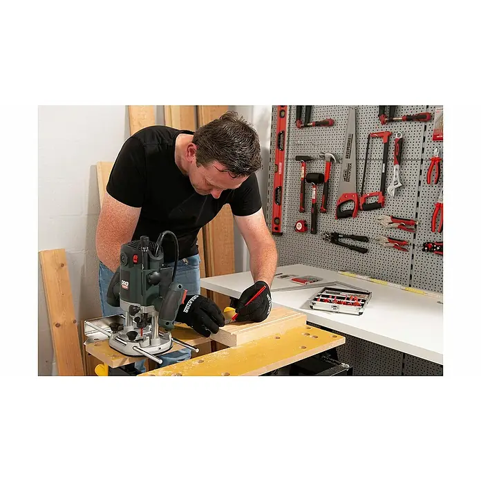 PowerPlus Fresadora de superficie POWP6050 - 2.100 W, Carrera máx. de fresado: 60 mm - Un hombre marca madera con un lápiz junto a una fresadora para cortes precisos. El taller está equipado con herramientas.