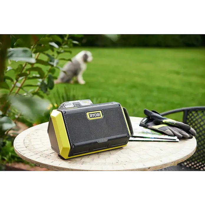 Ryobi ONE+ Altavoz Bluetooth RBT18-0 - 18 V, Sin batería, Potencia de salida altavoz: 2 x 10 W - Radio Ryobi ONE+ con batería sobre una mesa de jardín con guantes, con un perro al fondo sobre un césped verde.