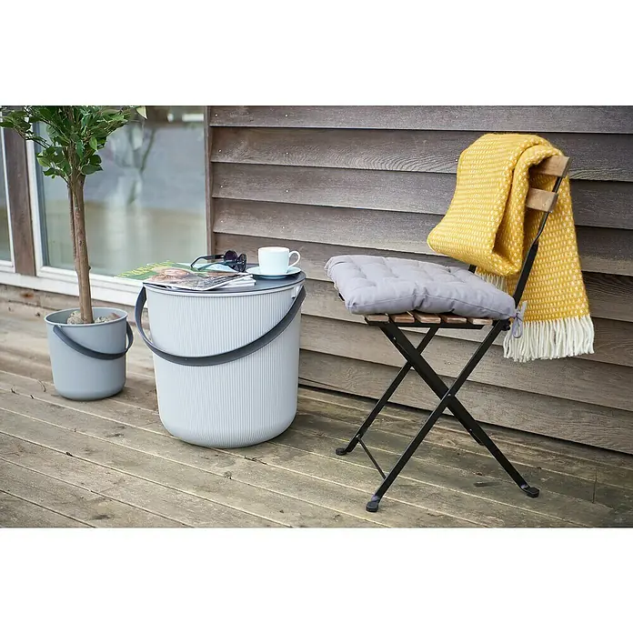 Eimer Akita - L x B x H: 480 x 446 x 445 mm, 53 l, Grau - Nahaufnahme von einem grauen Eimer-Tisch mit einem Getränk und Zeitschriften darauf neben einem Stuhl mit Kissen und gelber Decke.