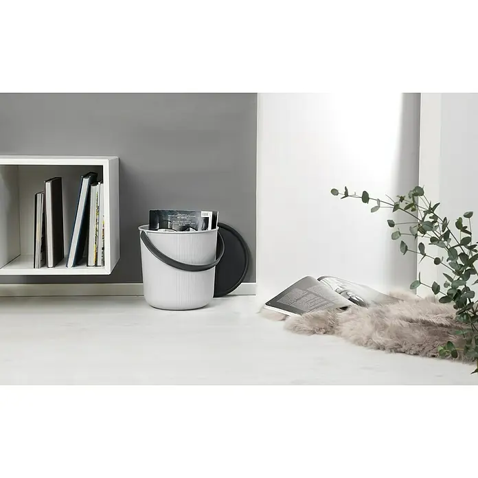 Eimer Akita - L x B x H: 277 x 271 x 258 mm, 10 l, Grau - Ein heller Eimer mit dunklem Griff und Deckel steht neben einem weißen Regal mit Büchern. Dekorative Elemente wie Pflanzen und Magazine ergänzen das Bild.