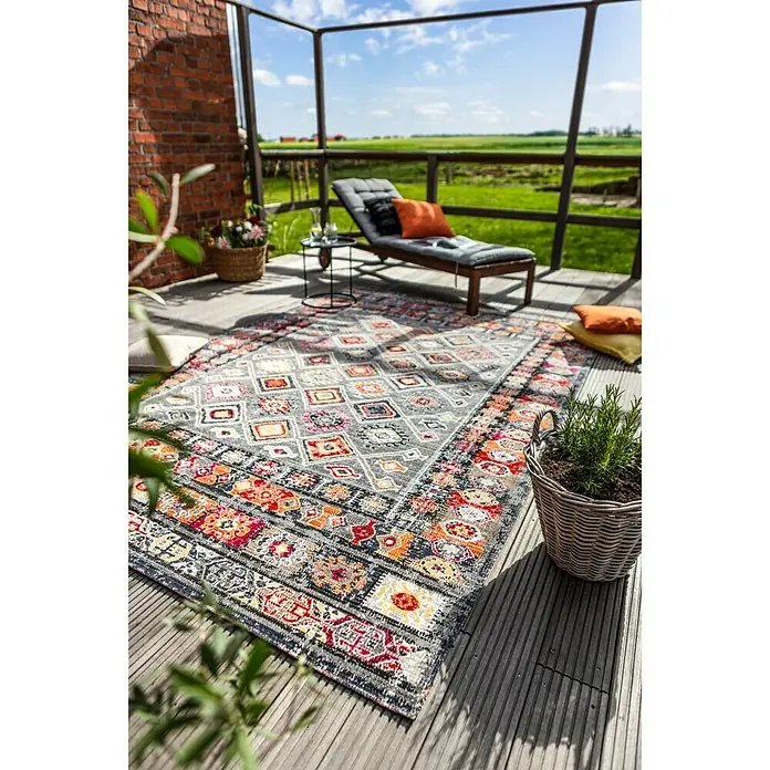 Outdoor-Teppich Bonnie - Bunt Hell, 230 x 160 cm, 100 % Polypropylen - Ein grauer Teppich mit bunten Mustern liegt auf einer Terrasse mit Liegestuhl und Pflanzenkörben im Hintergrund.