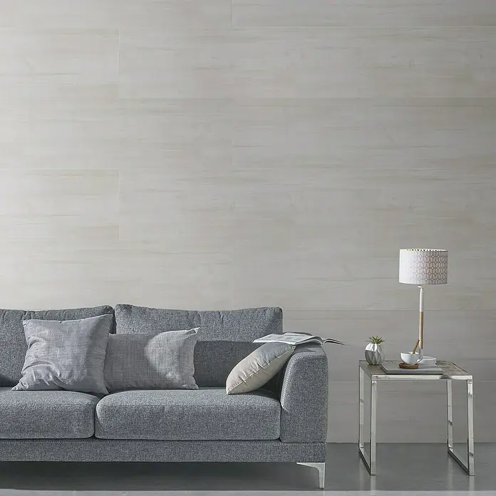 Grosfillex Panel de revestimiento Attitude Wood Beige - 260 cm x 37,5 cm x 8 mm - Un sofá gris con almohadas y una lámpara en una mesa auxiliar frente a una pared clara.