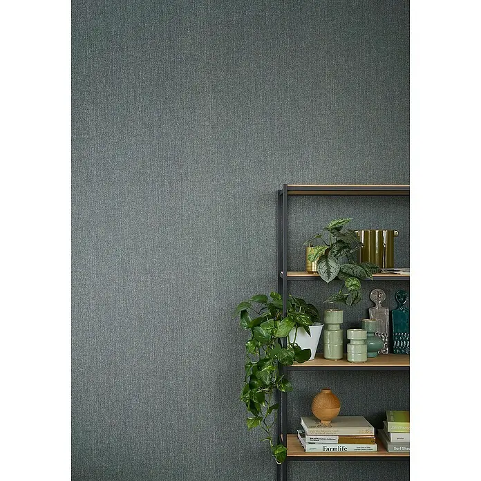 SCHÖNER WOHNEN-Kollektion Vliestapete Unistruktur - Grün/Grau, Uni, 10,05 x 0,53 m - Detailaufnahme eines Regals mit Zimmerpflanzen, Keramikvasen und Büchern vor einer grauen Textiltapete.