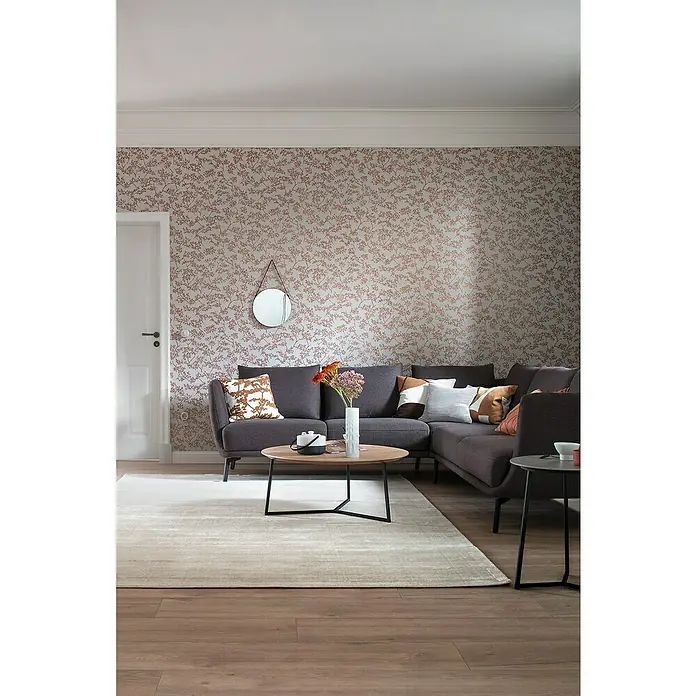 SCHÖNER WOHNEN-Kollektion New Spirit Vliestapete Struktur I - Grau/Gold, Floral, 10,05 x 0,53 m - Graues Ecksofa mit Zierkissen und einem runden Couchtisch auf einem hellen Teppich in einem gemütlichen Wohnzimmer mit floraler Tapete.