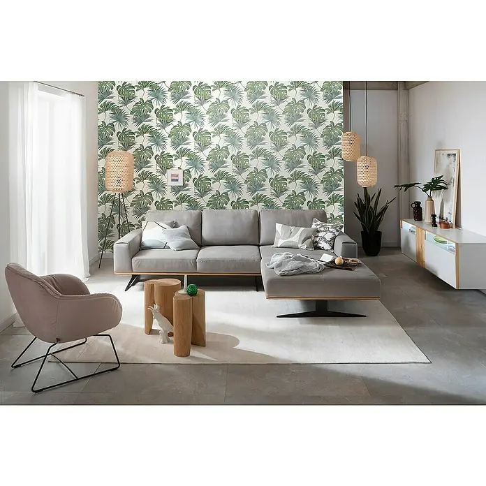 SCHÖNER WOHNEN-Kollektion New Spirit Vliestapete Fensterblatt - Grün/Weiß, Floral, 10,05 x 0,53 m - Ein modernes Wohnzimmer mit einem grauen Ecksofa, Holzhockern und einer gemusterten Tapete. Die sanfte Beleuchtung schafft eine einladende Atmosphäre.