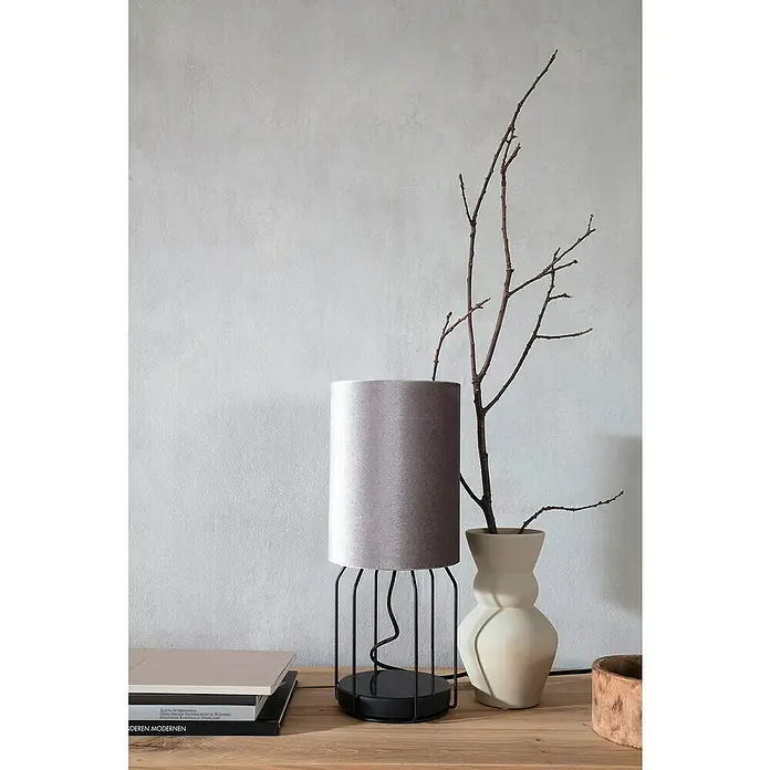 SCHÖNER WOHNEN-Kollektion New Spirit Vliestapete - Shadowgrau, Uni, 10,05 x 0,53 m - Elegante Stehleuchte mit grauem Schirm und filigranem Metallgestell neben einer Vase mit Zweigen auf einem Holztisch.