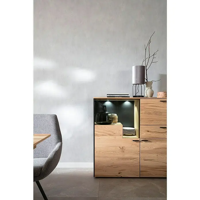 SCHÖNER WOHNEN-Kollektion New Spirit Vliestapete - Shadowgrau, Uni, 10,05 x 0,53 m - Nahaufnahme eines Sideboards aus Holz mit Fächern und Schubladen, dekoriert mit einer Lampe und Vasen.
