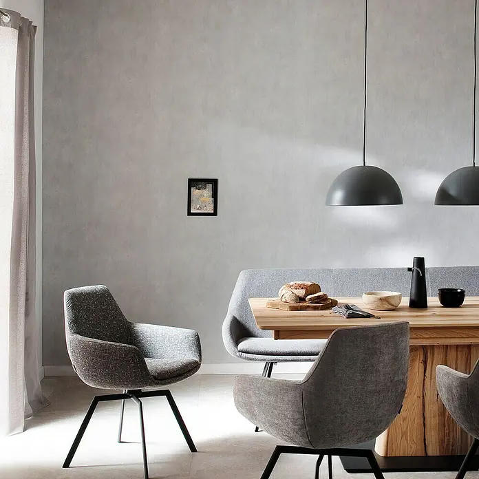 SCHÖNER WOHNEN-Kollektion New Spirit Vliestapete - Shadowgrau, Uni, 10,05 x 0,53 m - Esszimmer mit Holztisch, grauen Stühlen und Hängelampen. Die stilvolle Einrichtung schafft eine moderne und einladende Atmosphäre.